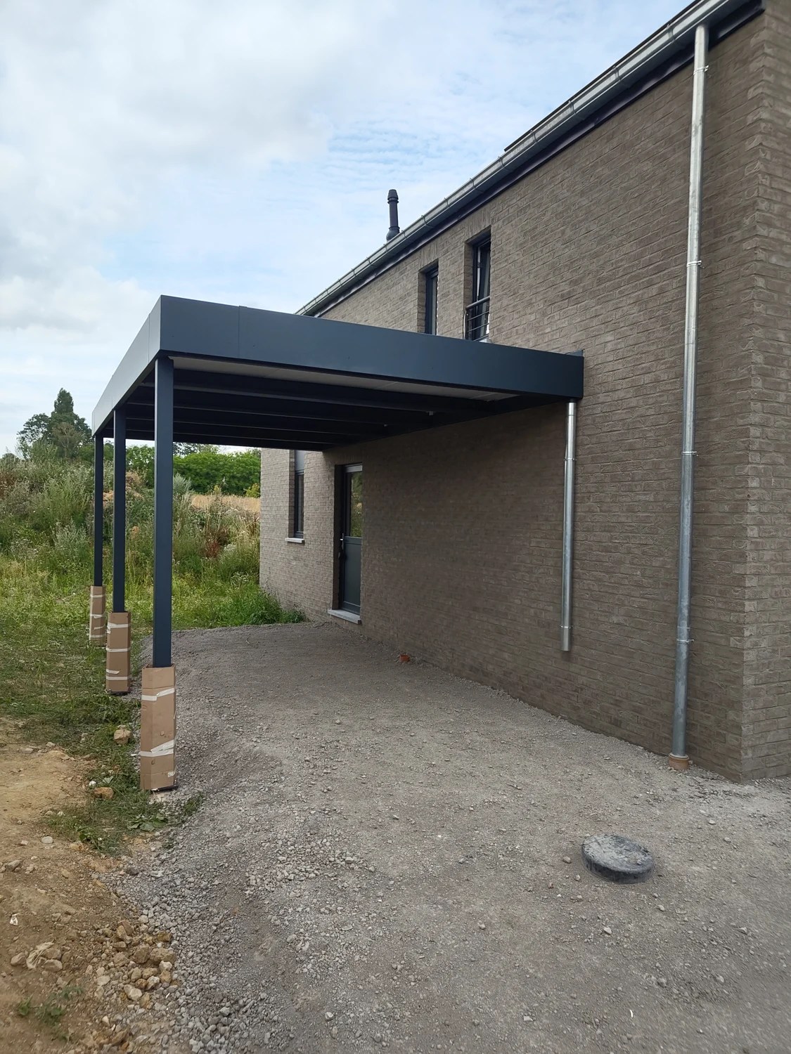 Carport adossé à une habitation en briques, avec poteaux métalliques noirs